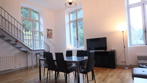 A modern living room with a dining area, large windows, a staircase, and a flat-screen TV. - Bright city-center loft, near thermal baths, for 4 guests (La Bourboule)