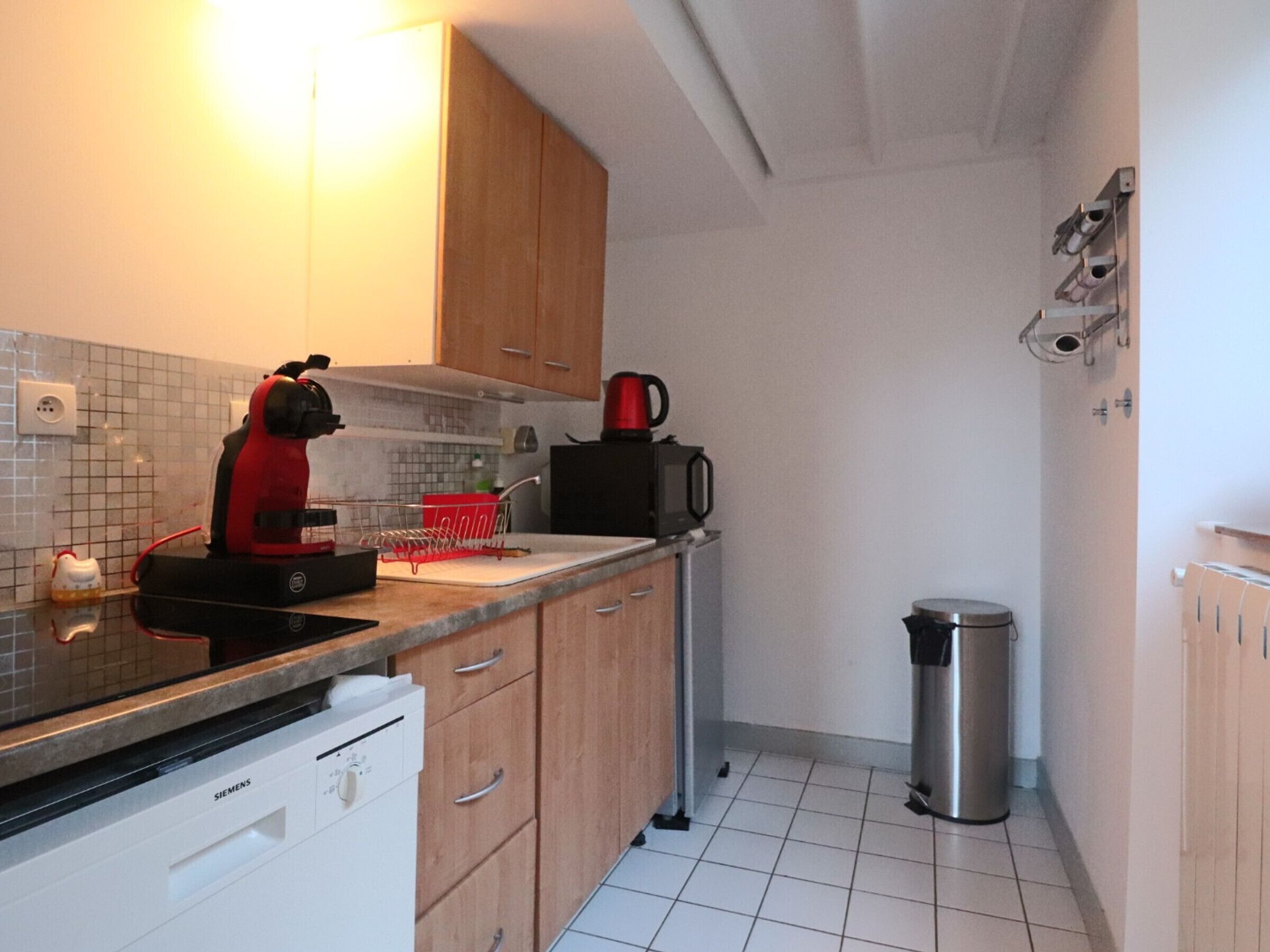 Eine kleine Küche mit Geschirrspüler, einer roten Kaffeemaschine, einer schwarzen Mikrowelle und einem Mülleimer.