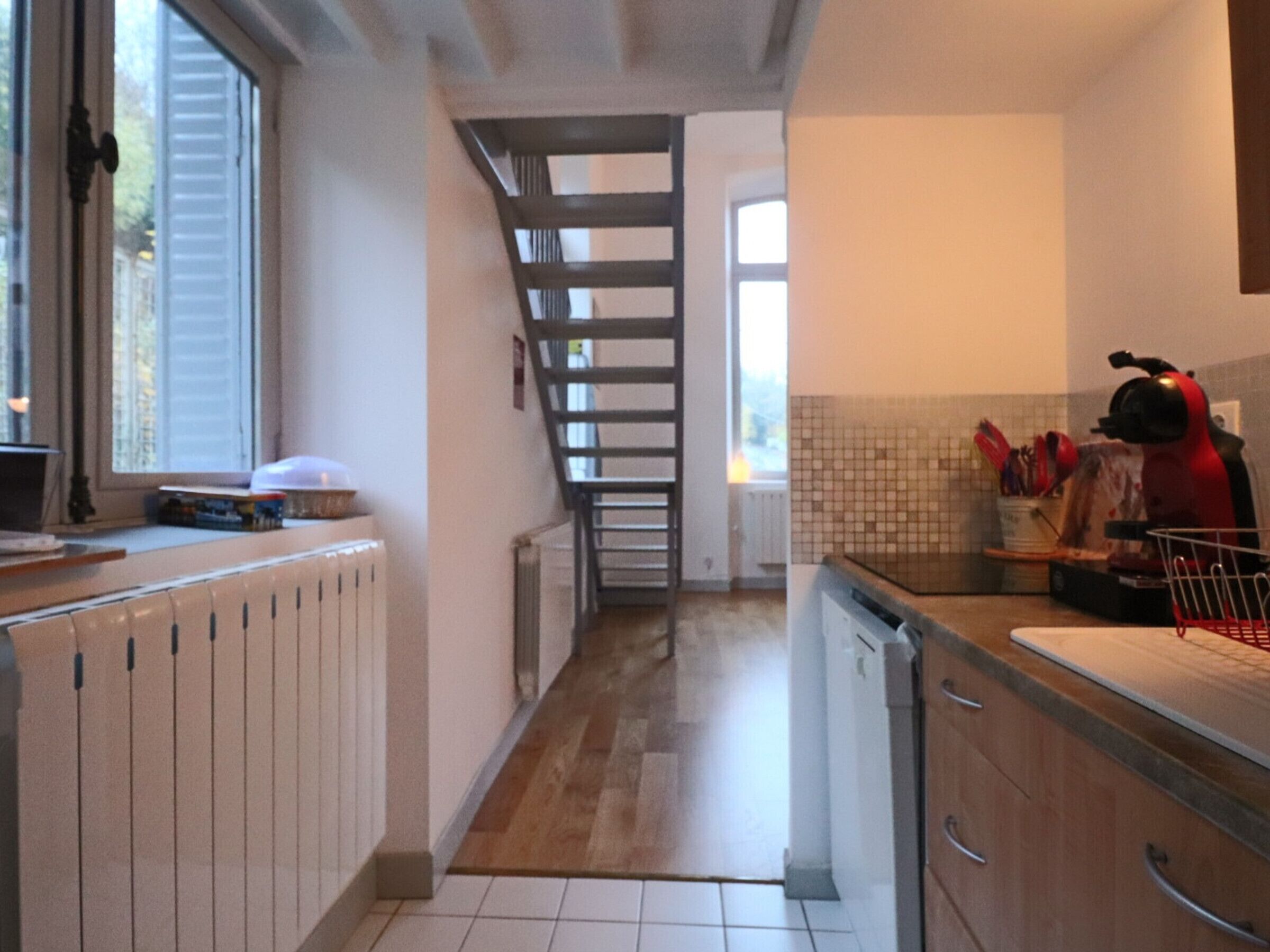 Una cocina estrecha con una escalera de madera, suelo de baldosas blancas y un electrodoméstico rojo sobre la encimera.