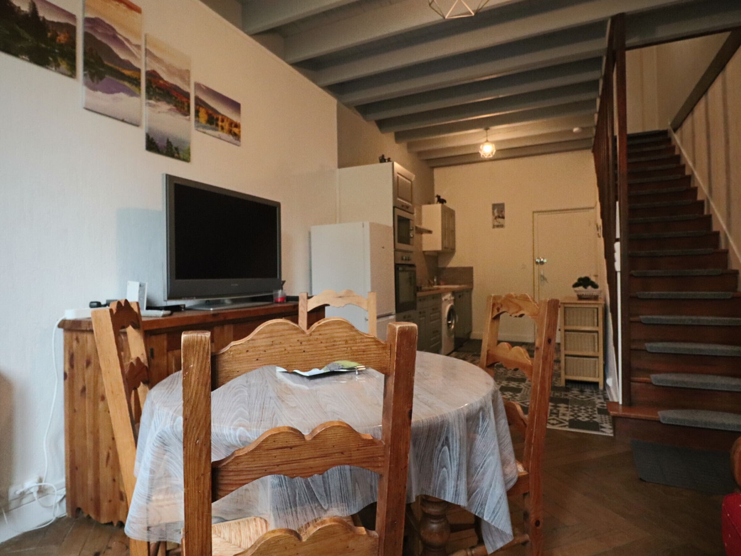 Uma área de jantar com uma mesa redonda e cadeiras de madeira, uma televisão em um suporte, uma cozinha ao fundo e uma escada que sobe.