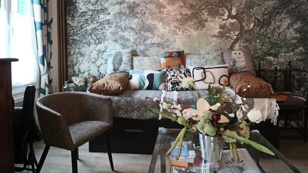 Een woonkamer met een bank, fauteuils en een salontafel. Er is een boekenkast en een muur met een bosreliëf.