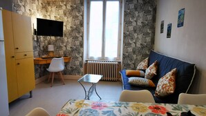 En kompakt opholdsstue med et skrivebord, en sofa, et spisebord og et fjernsyn ophængt på væggen.
