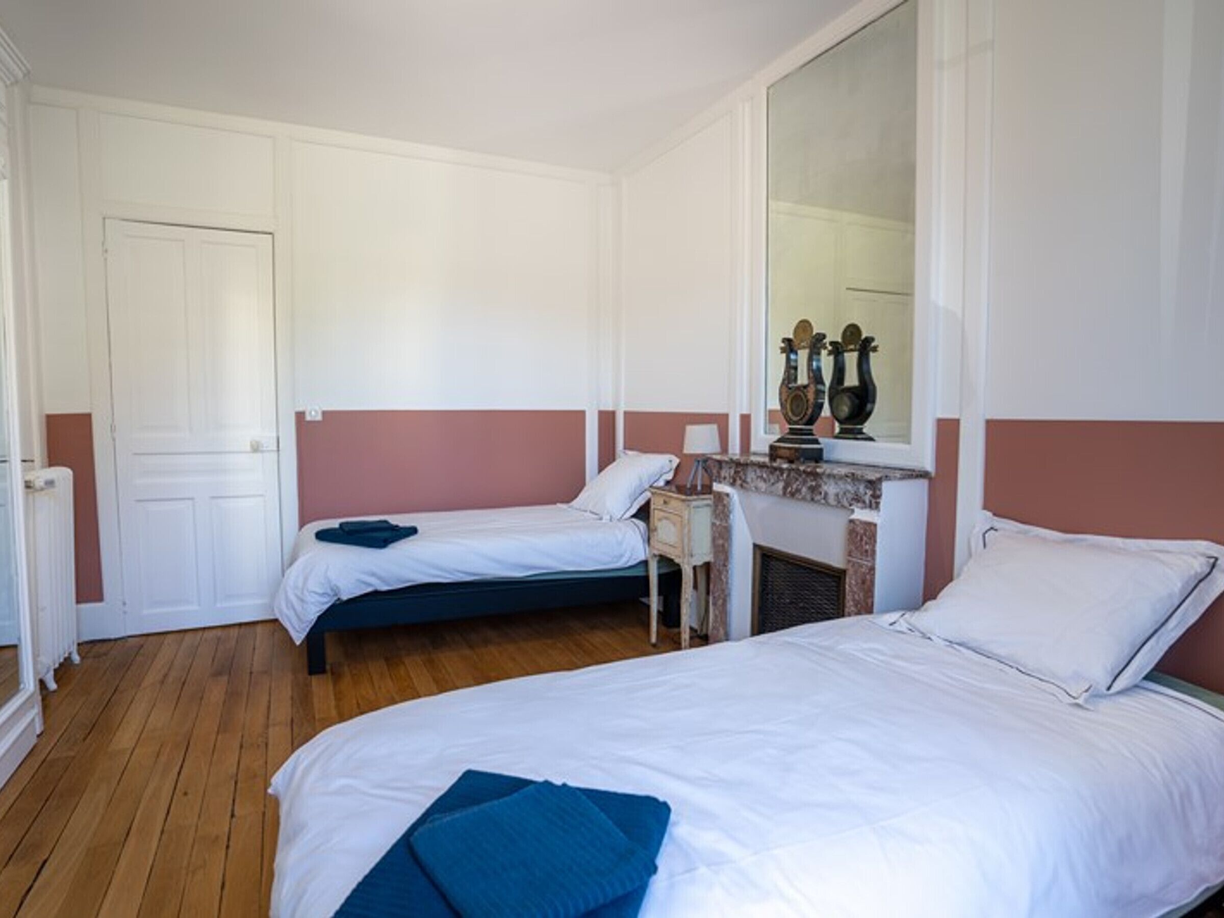 Una stanza con due letti singoli, pavimento in legno, una porta bianca e un camino con oggetti decorativi.