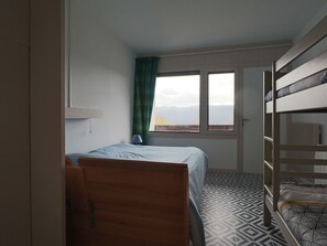 A room with a bunk bed, a single bed, a patterned floor, a window with a view, and a curtain. - Charming Duplex for 4 Pers. with Private Parking in Murat-le-Quaire (Murat-le-Quaire)