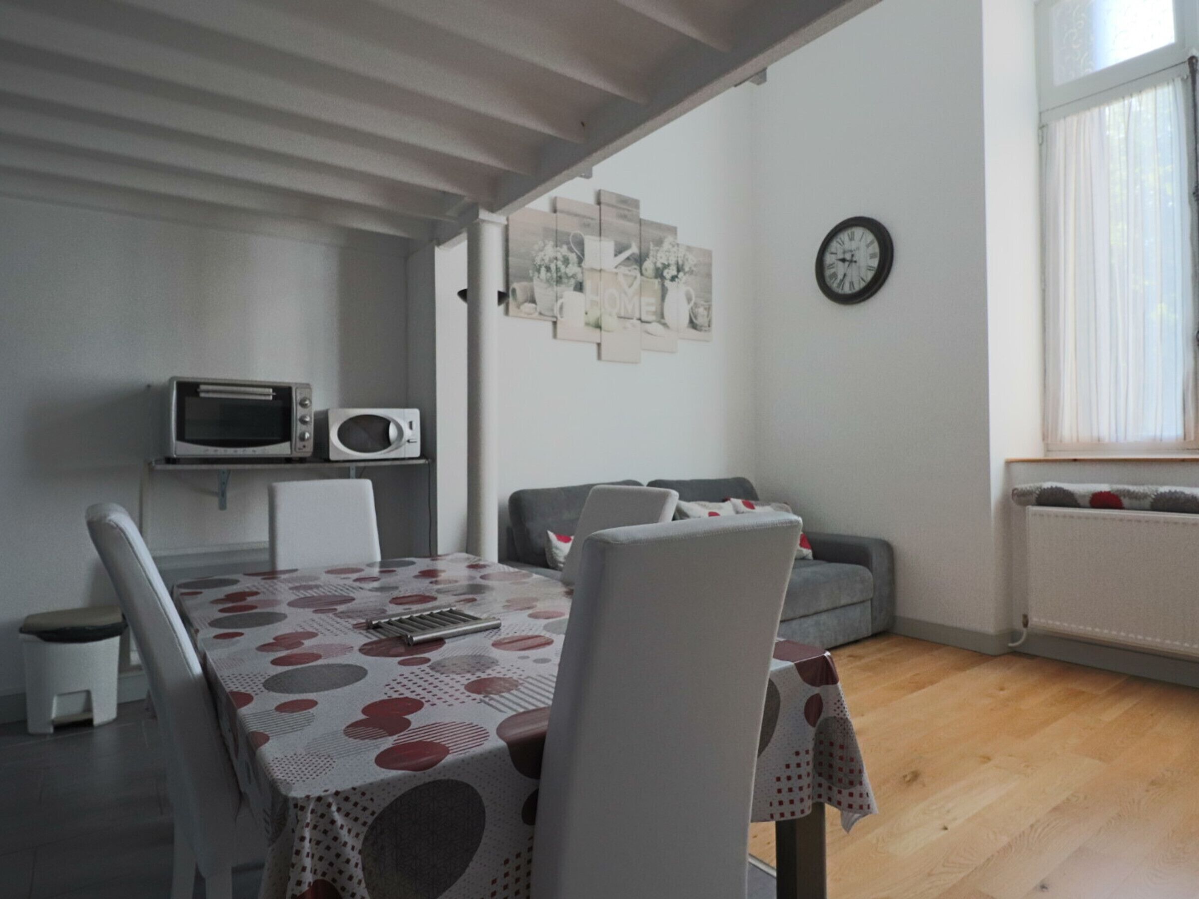 Uma área de refeições com uma mesa coberta por um guardanapo xadrez, duas cadeiras brancas, um micro-ondas em uma prateleira, um sofá cinza e um relógio de parede.