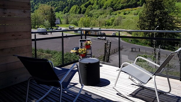 A balcony with two lounge chairs, a wicker table, and a view of a road with cars and a mountain in the background. - Apartment with large sunny terrace, close to spa and skiing (Mont-Dore)