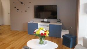 A modern living room with a large wall clock, a flat-screen TV, a blue entertainment unit, and a round white table with a vase of flowers. - Apartment with large sunny terrace, close to spa and skiing (Mont-Dore)
