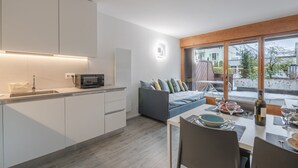 A modern kitchen with white cabinets, a microwave, and a sink. There is a dining area with a table set for two, a sofa, and a window with a view of a snowy landscape.