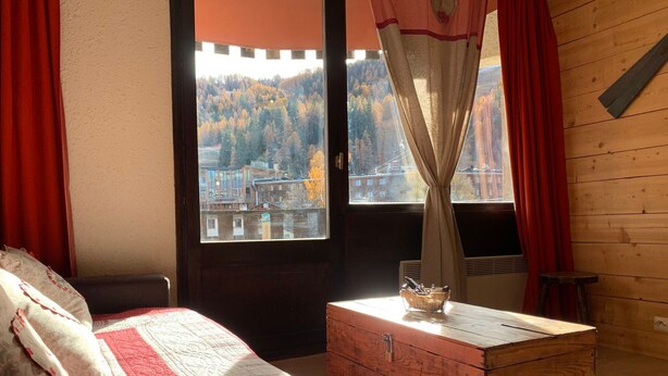 A bedroom with a bed, a wooden chest, and a window with a view of a forested area. - Bright studio in Pra-Loup for 4 pers, south balcony (Pra-Loup)