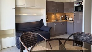 Ett kompakt kök med en inbyggd mikrovågsugn, en diskho och en kaffemaskin. I vardagsrummet finns en mörkfärgad soffa och fåtöljer i rotting.