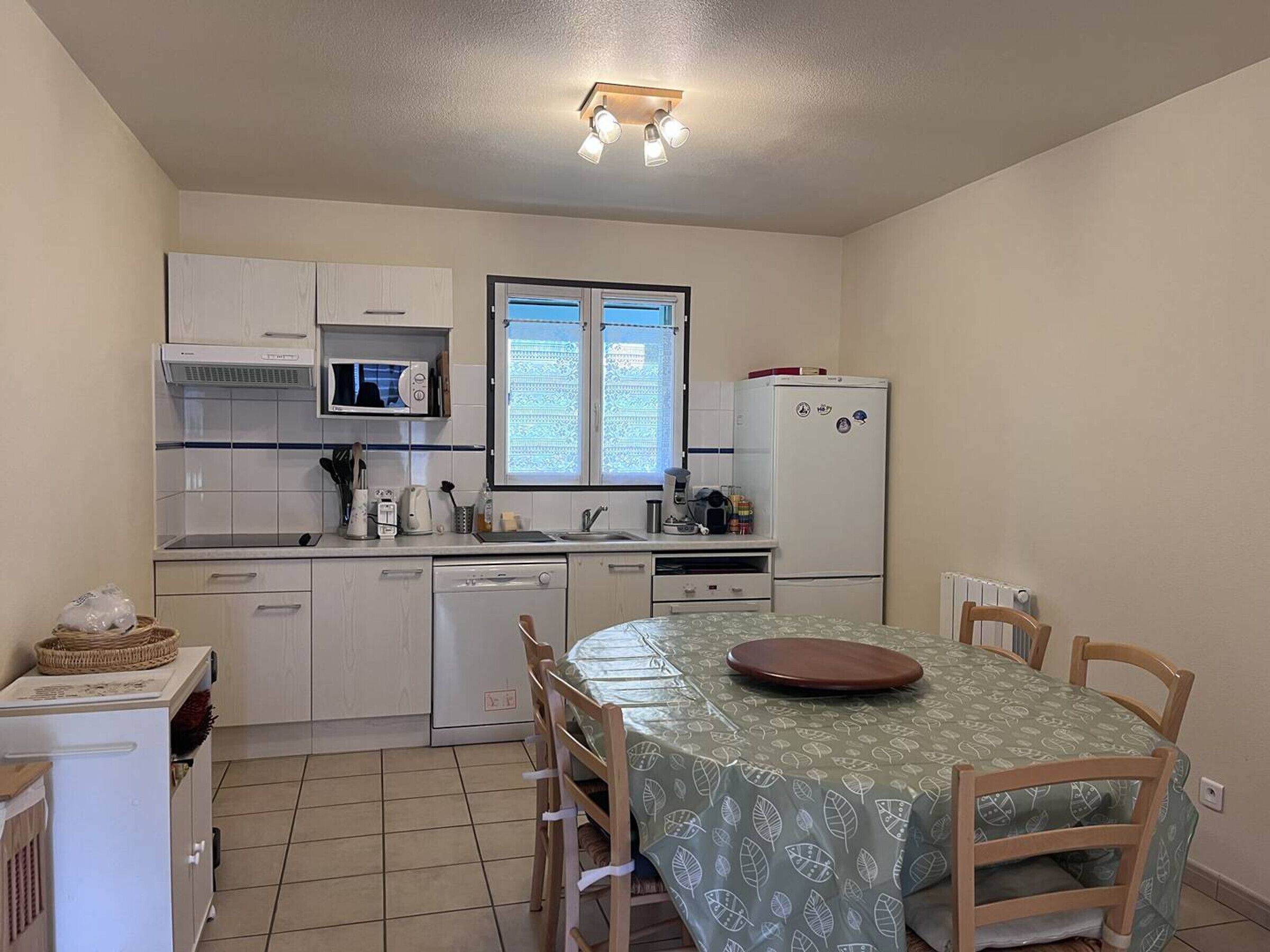 Une cuisine compacte avec des meubles blancs, un four à micro-ondes, un réfrigérateur et une fenêtre équipée de stores.