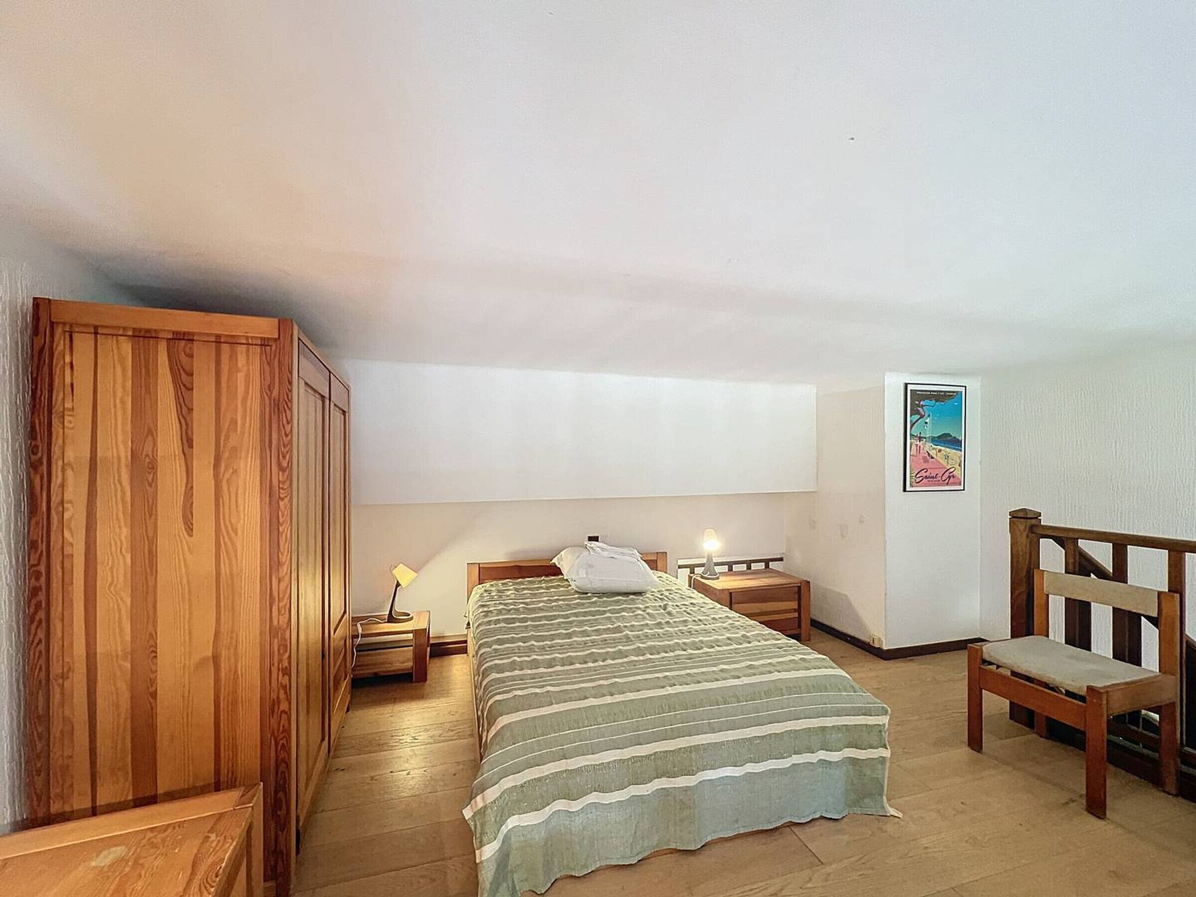 Un dormitorio con cama, armario de madera, mesitas de noche, una silla y un cartel en la pared.