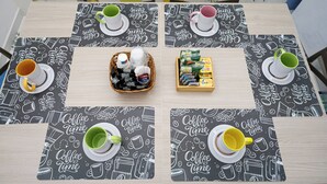 A dining table with four chairs, each with a coffee-themed placemat. There are four cups and saucers, one on each chair. A basket with various items is placed in the center. - Apartment Donn'Anna Int 14 (Napoli)
