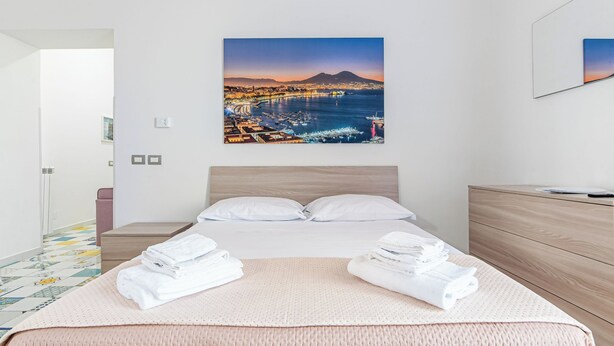 A modern hotel room with a bed, a wooden dresser, and a large cityscape painting on the wall. - Apartment Donn'Anna Int 14 (Napoli)