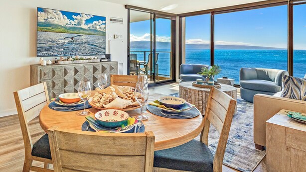 A dining area with a large table set for a meal, a view of the ocean through large windows, and a painting on the wall. - 1BD Peaceful Oceanfront Maui Escape w/ AC, Lanai & Island-to-Island Views - Mahana 1118 (Lahaina)