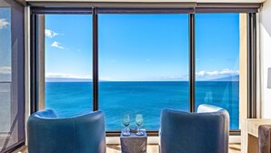 A room with a large window overlooking the sea, two chairs, a small table with two glasses, and a wooden floor. - 1BD Peaceful Oceanfront Maui Escape w/ AC, Lanai & Island-to-Island Views - Mahana 1118 (Lahaina)