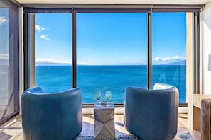 Ein Zimmer mit großem Fenster mit Meerblick, zwei Stühle, ein kleiner Tisch mit zwei Gläsern und ein Holzboden.