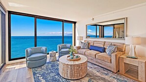 A modern living room with a large window overlooking the ocean, a round coffee table, a sofa, and a side table. - 1BD Peaceful Oceanfront Maui Escape w/ AC, Lanai & Island-to-Island Views - Mahana 1118 (Lahaina)