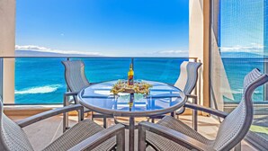 A balcony with a glass table, wicker chairs, and a bottle of wine. - 1BD Peaceful Oceanfront Maui Escape w/ AC, Lanai & Island-to-Island Views - Mahana 1118 (Lahaina)