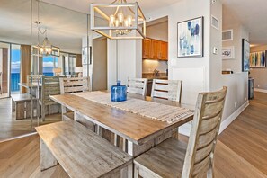Een ruime eethoek met een houten tafel en banken, een kroonluchter, en uitzicht op de keuken en woonkamer.
