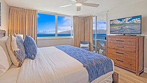 Ett hotellrum med en stor säng, en takfläkt, en byrå i trä och ett stort fönster med utsikt över havet.