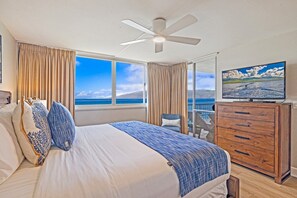 Hotelkamer met een groot bed, een plafondventilator, een houten dressoir en een groot raam met uitzicht op zee.