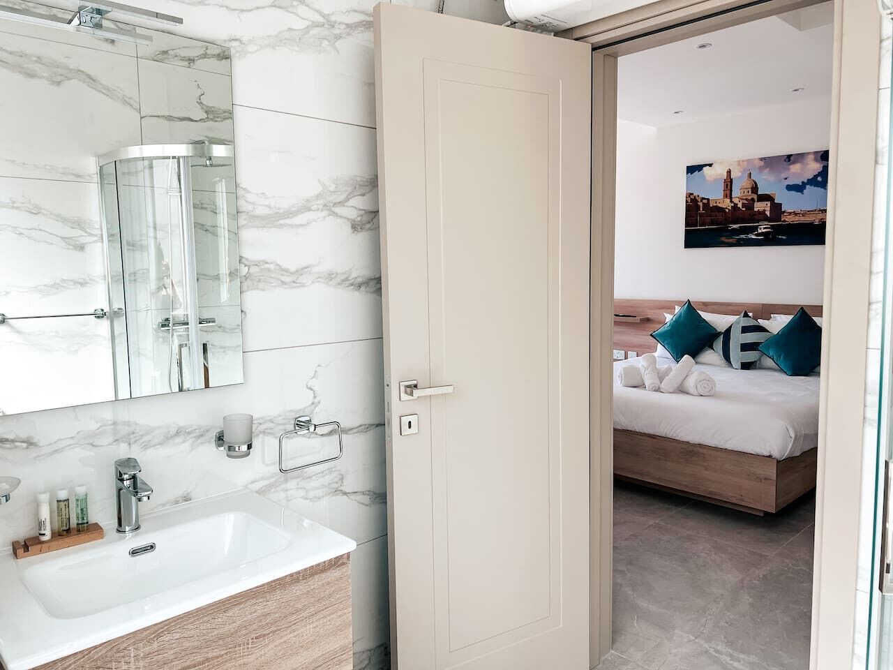 A modern bathroom with a marble vanity, a glass shower enclosure, and a bed with blue pillows in the adjacent room.