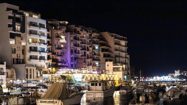 A marina with boats docked, a brightly lit building with balconies, and a night-time cityscape in the background. - Modern Escape Villa with Pool by ArcoCollection (St. Julian`s)