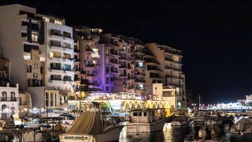 A marina with boats docked, a brightly lit building with balconies, and a night-time cityscape in the background.