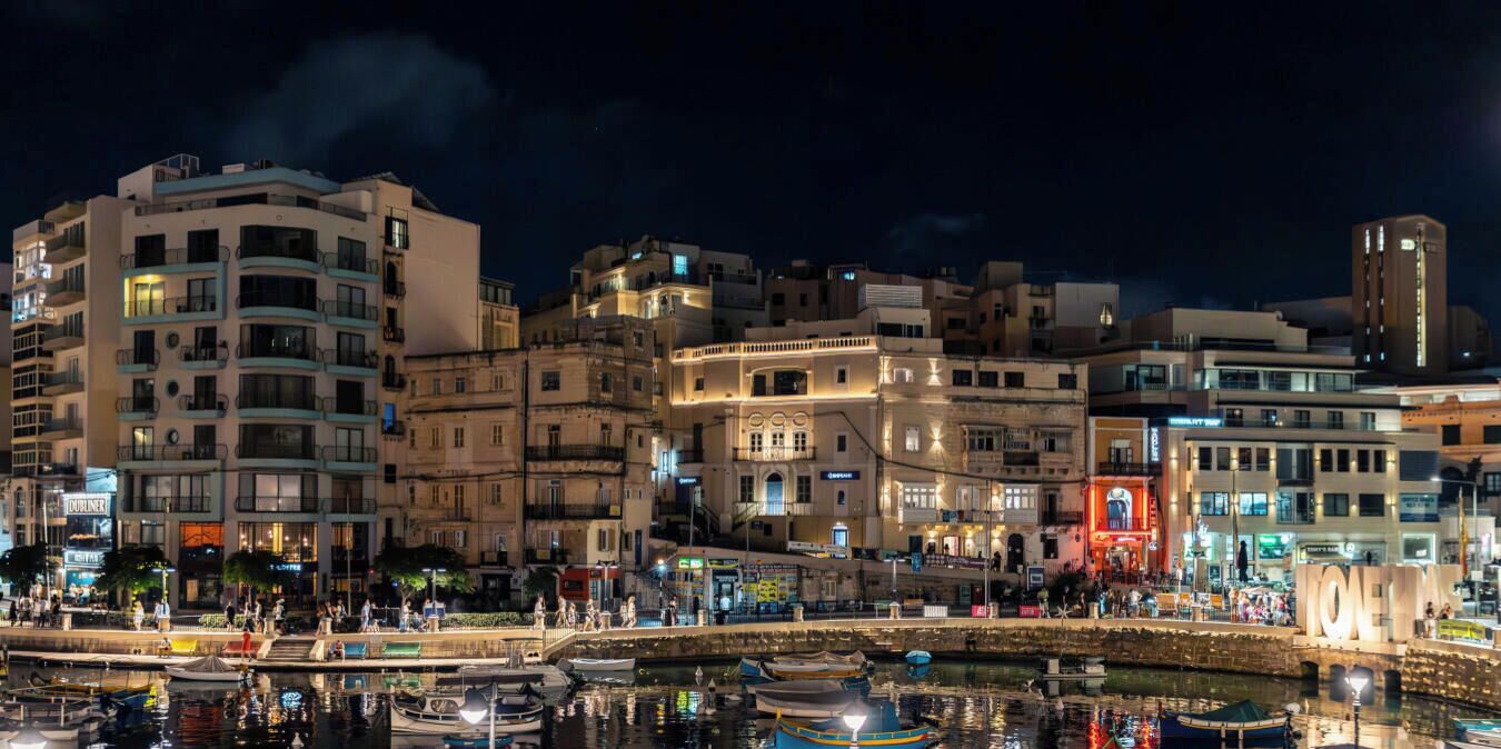 Paesaggio urbano con edifici, un molo con barche e una passeggiata pedonale.