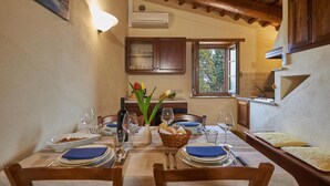 Un coin repas avec une table dressée pour quatre personnes, une fenêtre donnant sur un paysage verdoyant et un espace cuisine avec des placards en bois et un luminaire suspendu.