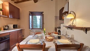 Un coin repas avec une table dressée pour deux personnes, des chaises en bois, une fenêtre avec vue et un espace cuisine équipé de placards et d’un four à micro-ondes.