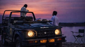 En Maruti-bil med to personer, en på taget og en indeni, parkeret ved vandkanten i skumringen.