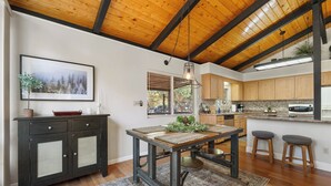 A modern kitchen with wooden beams, a dining table with stools, and a view of the outdoors.