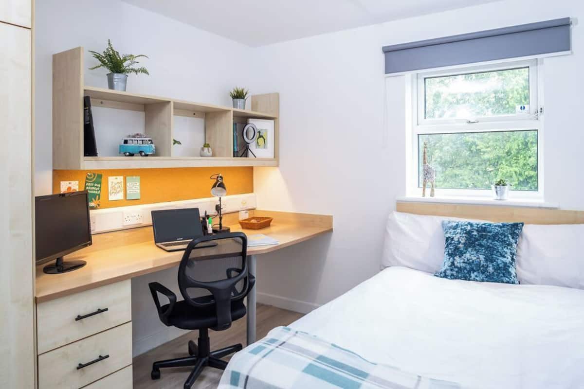 A bedroom with a bed, a desk with a computer, a chair, and shelves with plants and decorative items.