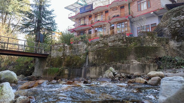A hotel with a balcony overlooking a river, a stone wall, and a bridge. - Elysian heights (Manali)
