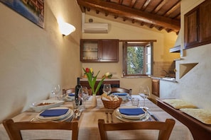 Un coin repas avec une table dressée pour quatre personnes, une fenêtre donnant sur un paysage verdoyant et un espace cuisine avec des placards en bois et un luminaire suspendu.