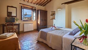 A bedroom with a bed, a desk, a chair, a radiator, a window with a view, and a vase with flowers. - Agriturismo Bonello Flat Standard Apartment with Pool, Shared Garden and Wi-Fi (Pienza)