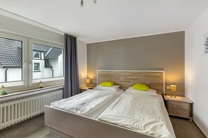 A double bed with white bedding, a wooden headboard, two bedside lamps, a nightstand, a window with curtains, and a radiator. - Room '1_Double Room No. 1' with Wi-Fi (Neuenrade)
