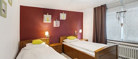 A room with two wooden beds, a red wall, framed pictures, and a window with blinds.