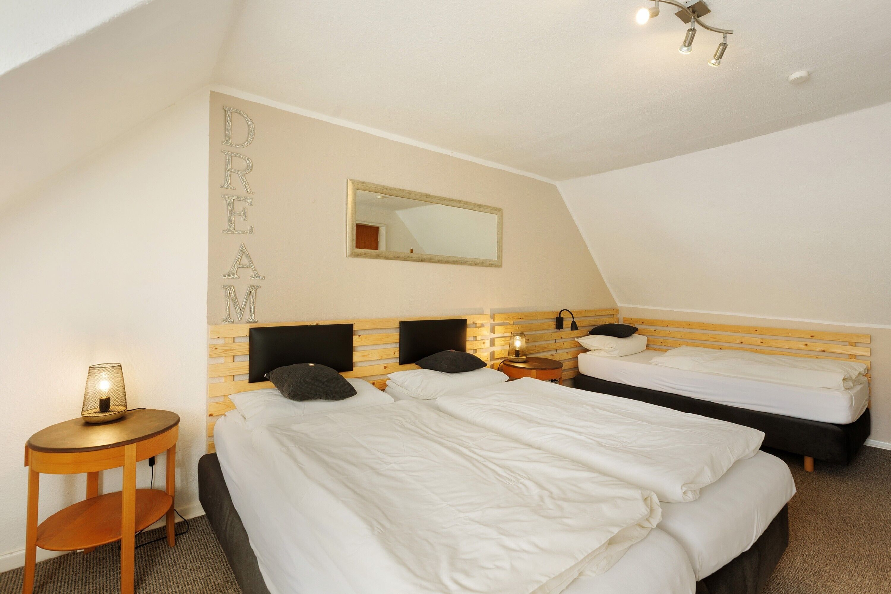 A hotel room with two beds, a bedside table with a lamp, a mirror on the wall, and a ceiling light fixture.