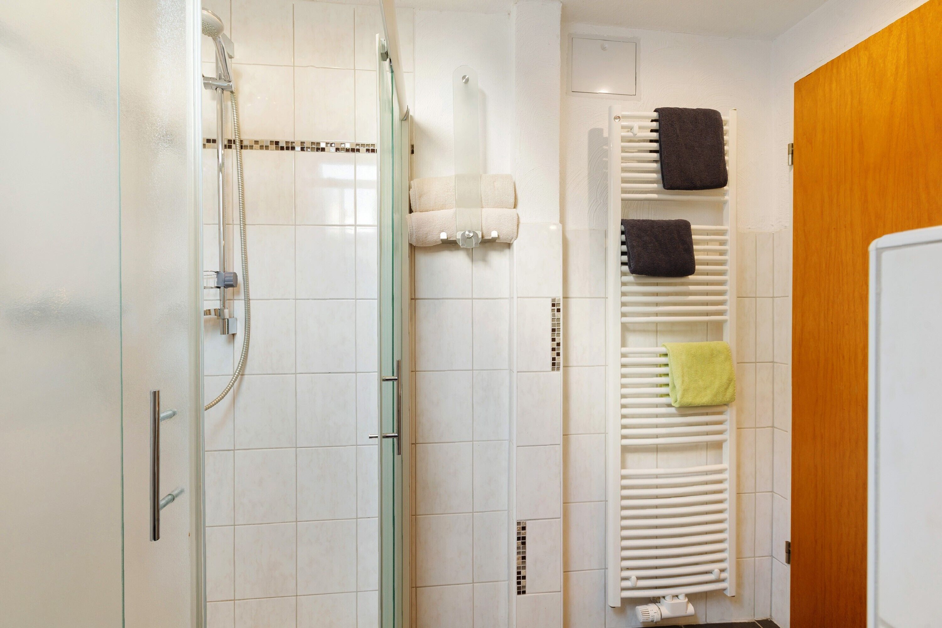Een badkamer met een douchecabine van glas, een witte handdoekhouder met meerdere handdoeken, en een houten deur.