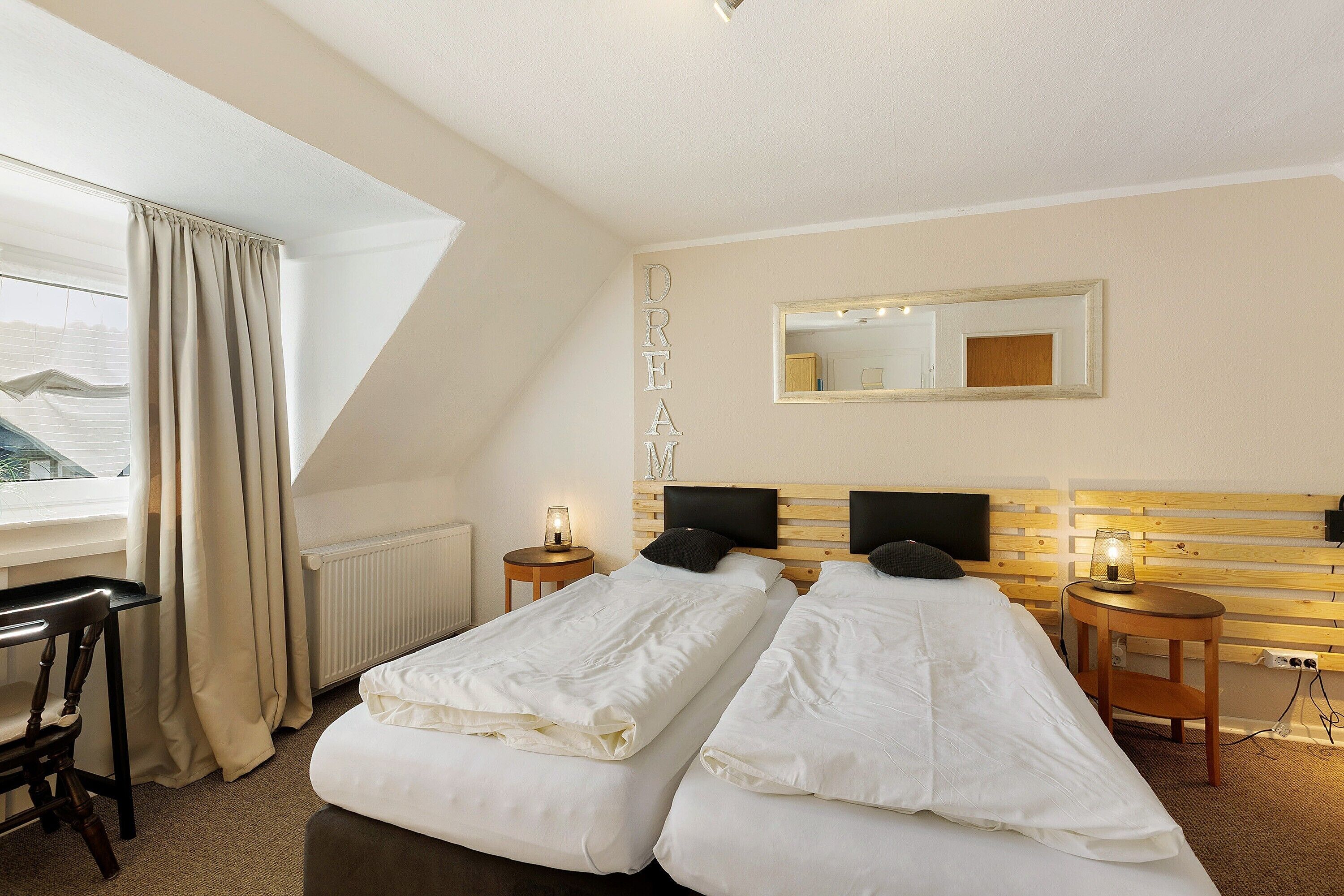 A hotel room with two beds, a desk, a chair, a window with curtains, a bedside table with a lamp, a mirror, and a radiator.