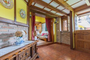 Un interior rústico con pared de ladrillo, vigas de madera, suelo de baldosas, un banco de madera con un jarrón y una puerta que da a otra habitación.