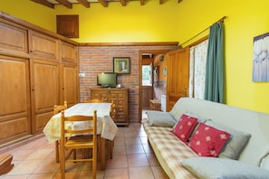 A dining area with a table, chairs, and a television. There is a sofa with cushions and a window with curtains.