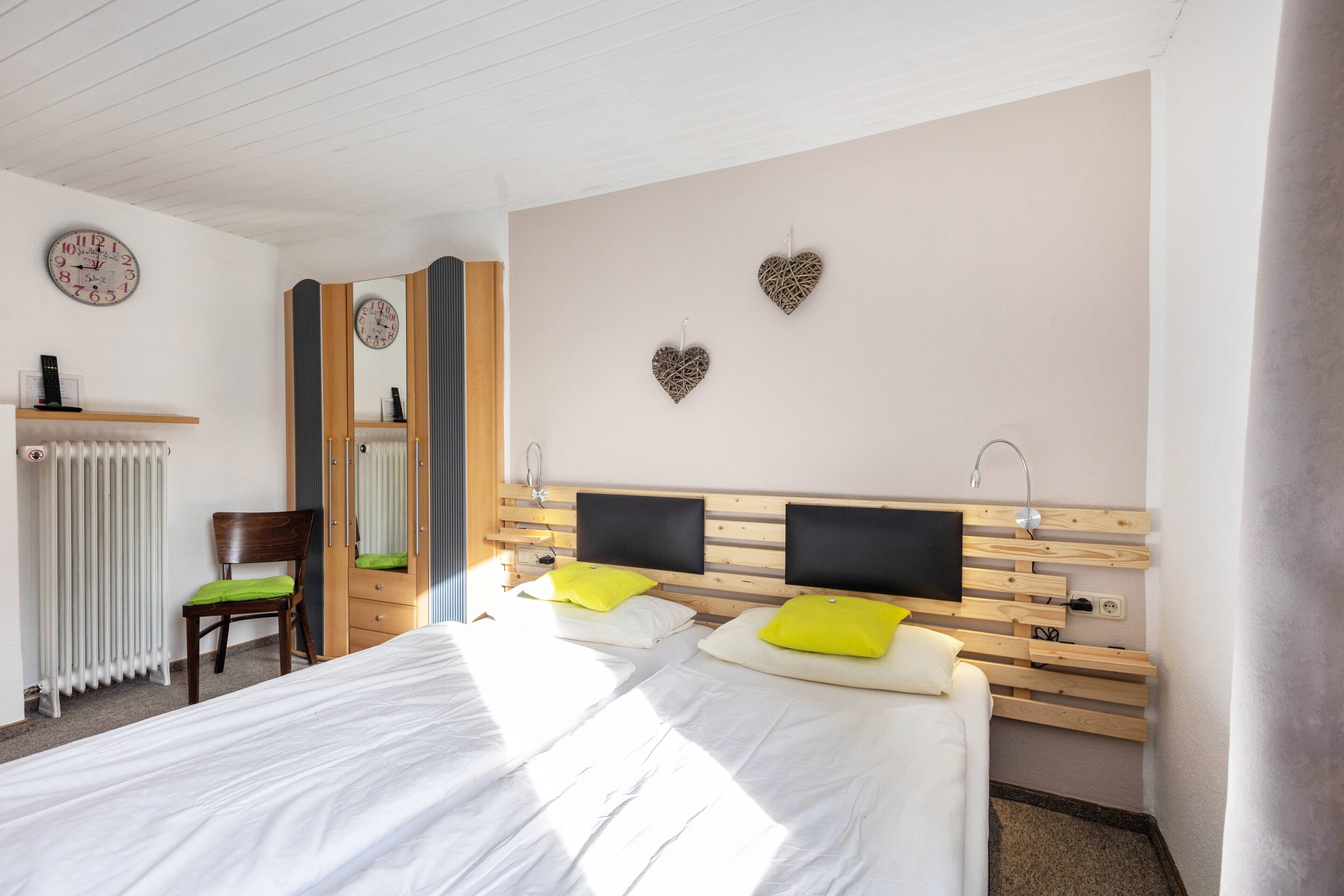 A hotel room with a large bed, two bedside tables, a chair, a mirror, a clock, and decorative heart-shaped wall art.
