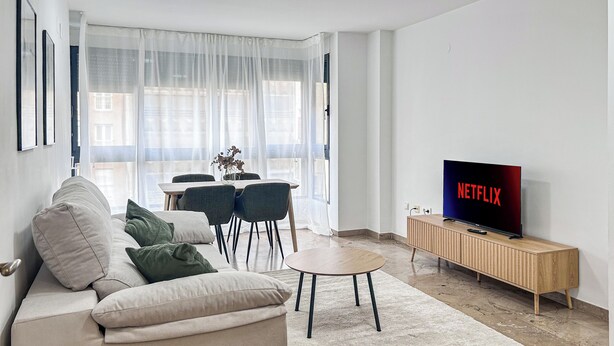 A modern living room with a sofa, a dining table with chairs, a TV on a wooden stand, and a large window with sheer curtains. - Nomads' Triangle" Central Park - Ruzafa - Turia (València)