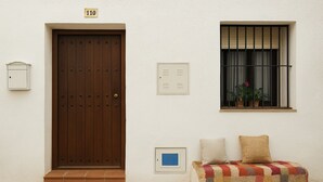 A room with a wooden door, a window with bars, a bench with cushions, and a small wall-mounted light fixture. - Country house in the Sierra Nevada- Almería. Nature, hiking and relaxation (Abrucena)