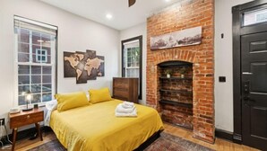 A bedroom with a brick fireplace, a bed with yellow bedding, a wooden dresser, and a large window. - Spacious, Historic 4Br Logan Circle Townhome (Washington D.C.)