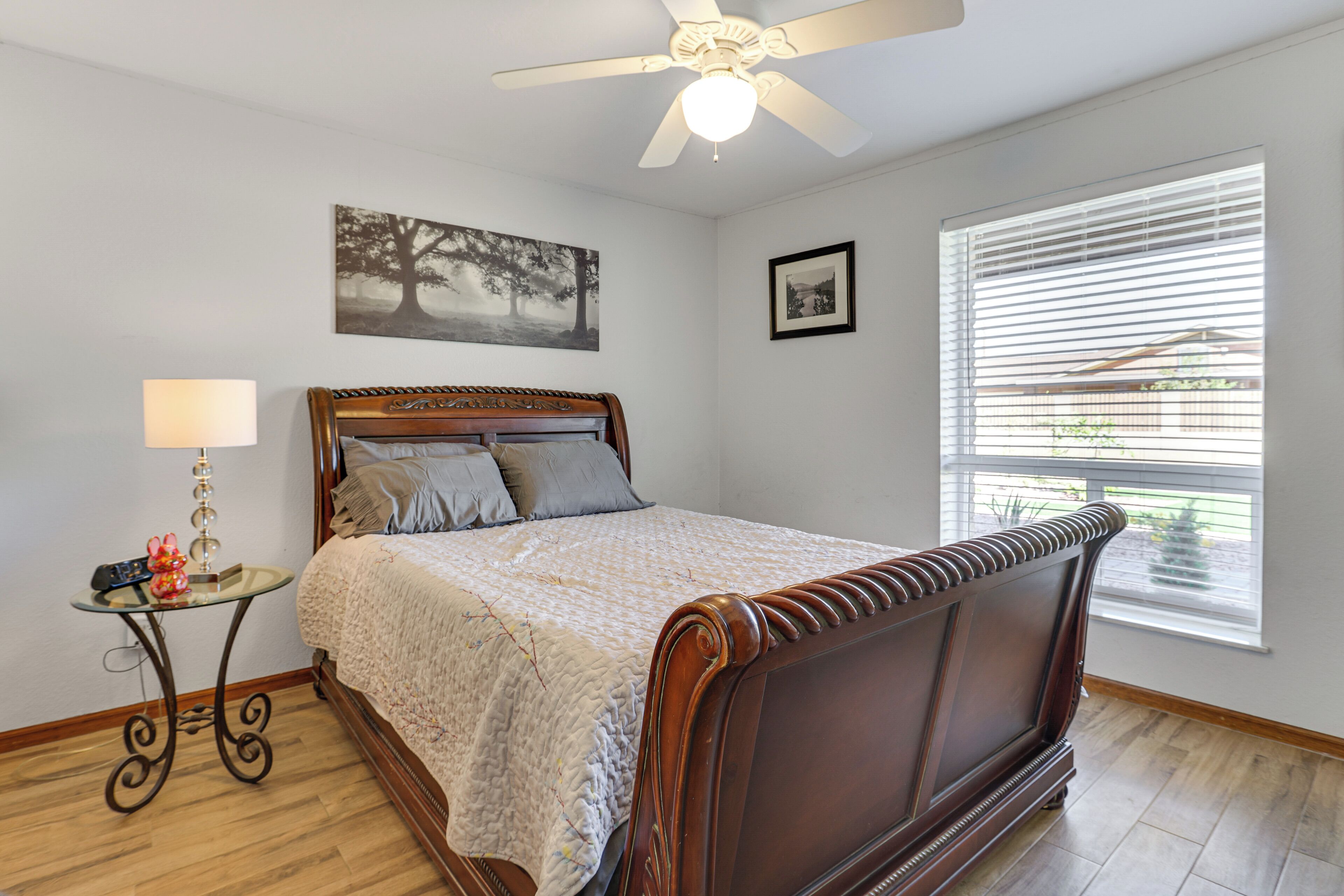 A bedroom with a wooden bed, a ceiling fan, a window with blinds, and a small table with a lamp.